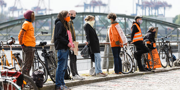 Menschen mit Atemschutzmasken und leuchtend orangefarbenen Kelidungsstücken sowie Transparenten stehen an einem Geländer, im Hintergrund Hafenkräne