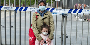 eine Mutter mit ihrem Kind vor einem Zaun, der mit Polizeiflatterband umwickelt ist, beide tragen Mundschutz
