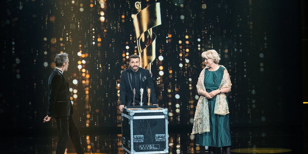 drei Menschen stehen nebeneinander, im Hintergrund ein übergroßer goldener Filmpreis