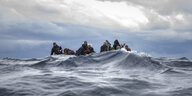 eine Welle verdeckt zur Hälfte kleines Boot, in dem viele Männer sitzen