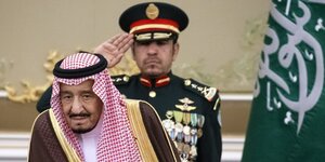 Saudi-Arabiens König Salman mit traditioneller Kopfbedeckung. Hinter ihm salutiert ein Militär