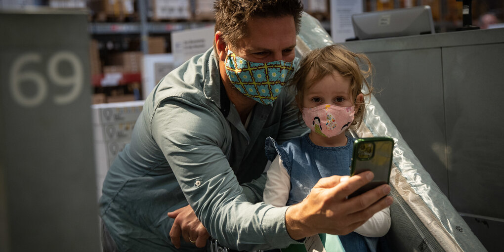 Vater und Tochter im Einkaufwagen, beide tragen Masken