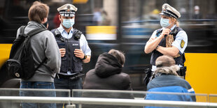 Zwei Polizeibeamte stehen vor drei Personen die auf der Bank vor einer Straßenbahnhaltestelle sitzen