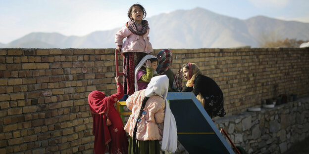 Kinder spielen neben einer Mauer auf einer alten Rutsche, im Hintergrund sind Berge
