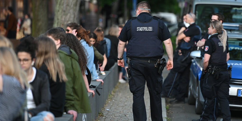 Menschen sitzen auf Bänken an einem Platz, die Polizeit patrouilliert.