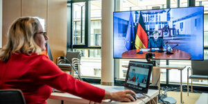 eine Frau mit Laptop auf dem Tisch, schaut Merkel im Fernsehen bei einer Rede zu