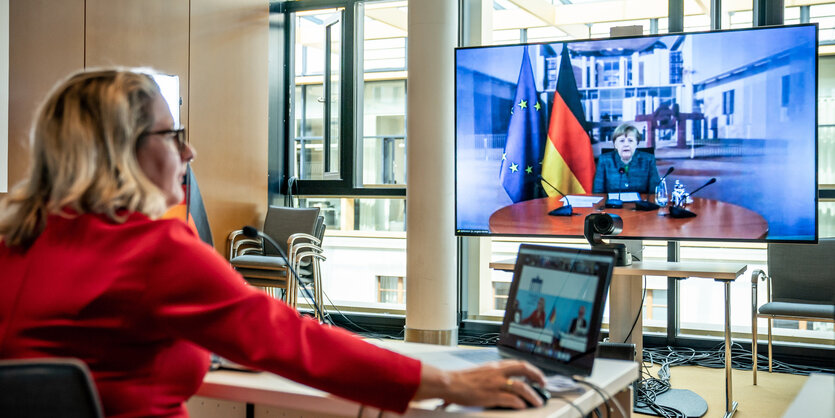 eine Frau mit Laptop auf dem Tisch, schaut Merkel im Fernsehen bei einer Rede zu