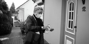 Eine Frau mit Mundschutz desinfiziert sich vor einer Haustür die Hände. Sie trägt Mundschutz.