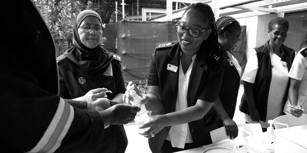 Im Milpark Hospital in Johannesburg, Südafrika, verteilt eine Mitarbeiterin handdesinfektionsmittel an andere Menschen. Sie drückt auf den Spender, während die anderen ihre Hände darunter halten. Die Frau lächelt.