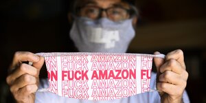 Ein Mann hält eine Gesichtsmaske in die Kamera auf der steht: Fuck Amazon