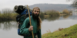 Mann mit Rucksack und Wanderstab steht am Flussufer