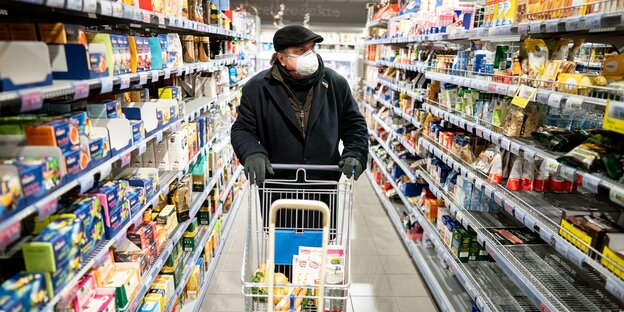 Links und rechts volle Regale: Ein Kunde kauft mit Mundschutz und Handschuhen am frühen Morgen in einem Supermarkt im Berliner Stadtteil Friedenau ein