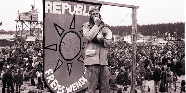 Ein Mann steht mit einem Megaphon vor einem Banner, auf dem steht "Republik Freies Wendland"
