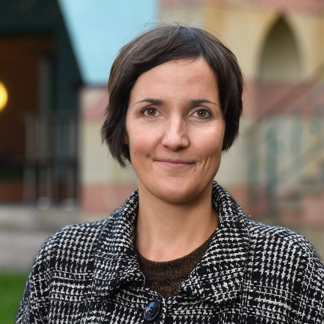 Tine Hanrieder, eine Frau mit mittellangen dunklen haaren. Sie trägt eine karierte Jacke mit Kragen und lächelt verschmitzt. Im Hintergrund steht ein Gebäude mit Bogen.