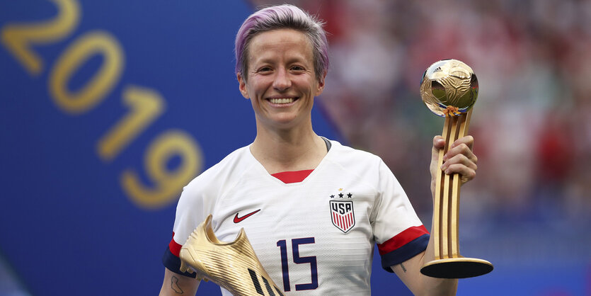 Megan Rapinoe lächelt, hat grau-violette Haare und hält stolz einen Pokal in die Höhe