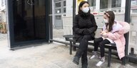Eine junge Frau und ihre kleine Tochter sitzen mit Mund-Nase-Schutz auf einer Bank an einer Tram-Haltestelle