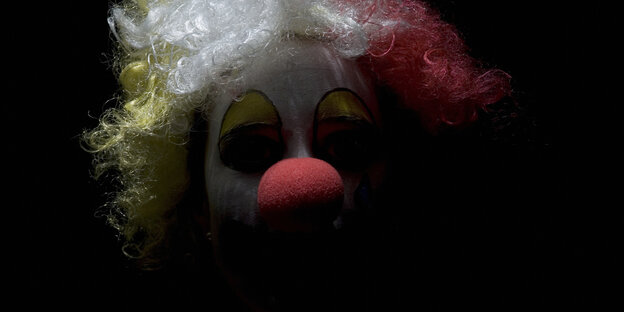 Ein Clown im Dunkeln, man sieht vor allem die rote Nase und die weiß-gelb-rote Perrücke