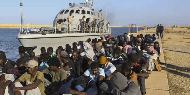 Schwarze Menschen sitzen auf einem Kai, im Hintergrund ein Boot der Libyschen Küstenwache.