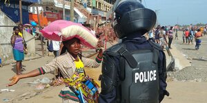 Eine uniformierte Polizeieinheit mit Helm und Rüstung steht vor einer eingeschüchterten jungen Frau, fir einen Sack auf dem Kopf trägt