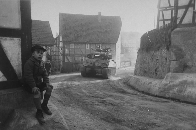 Ein Junge sitzt an einem Fachwerkhaus, während ein Panzer durch den Ort fährt