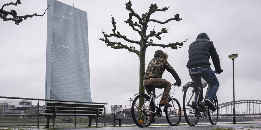 Zwei Radfahrer rollen im Regen am Main entlang, Dahinter ist die Zentrale der Europäischen Zentralbank