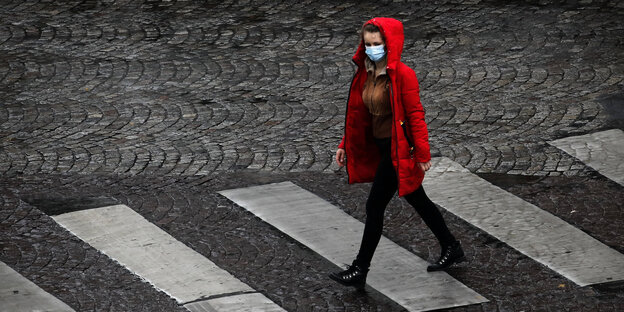 eine Frau in geht in einem roten Manel und Mundschutz über einen Zebrastreifen