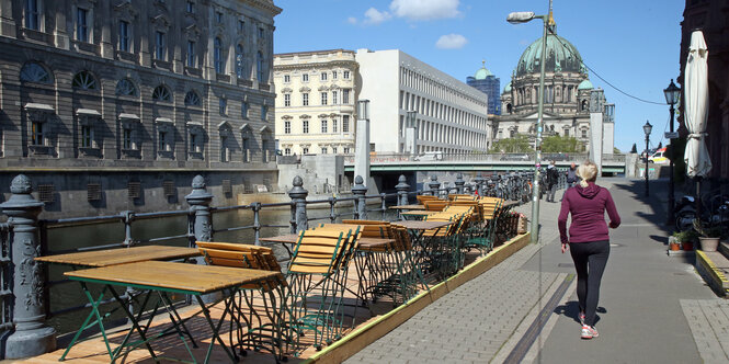 Eine Frau geht im Nikolaiviertel an leeren Tischen und Stühlen eines geschlossenen Restaurants vorbei. Im Hintergrund der Berliner Dom und das Humboldt-Forum zu sehen