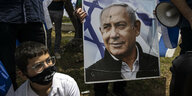 Ein Mann mit Mundschutz sitzt neben einem Plakat, das Benjamin Netanjahu zeigt.