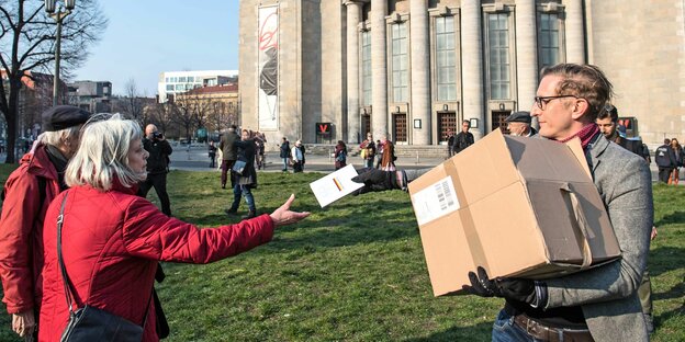 Anselm Lenz reicht einer Frau mit grauen Haaren ein Buch aus einer Kiste. Im Hintergrund steht die Schaubühne. Am Rosa-Luxemburg-Platz wurde die Hygiene-Demo abgehalten.