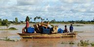 Drei Menschen transportieren auf einem Boot ihr Hab und Gut über einen über die Ufer getretenen Fluss