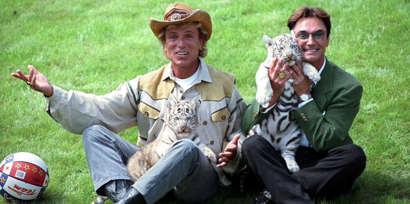 Siegfried trägt einen hellen Hut, Roy hat mittellange schwarze Haare, beide lachen und halten weiße Tiger in ihrem Schoß