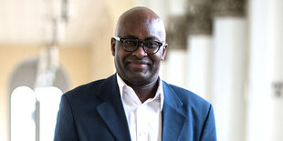 Achille Mbembe steht in der LMU München. Er ist ein älterer Mann ohne Haare und trägt über seinem weißen Shirt ein dunkelblaues Sakko. Er trägt eine runde Brille und lächelt.