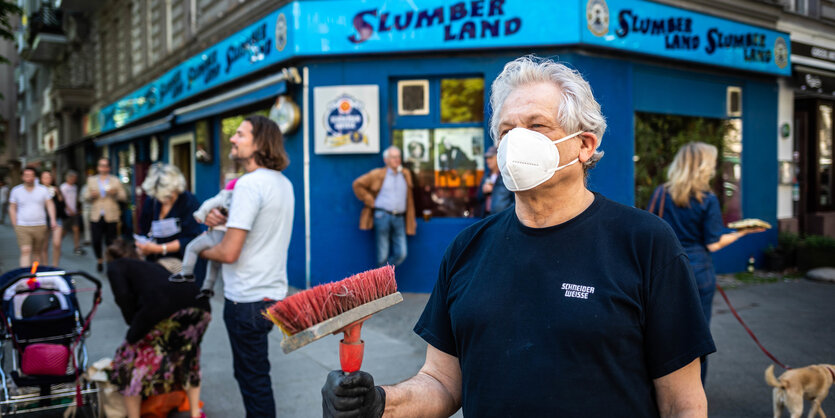 Wirt des Slumberland mit Besen und Mundschutz vor dem Lokal am Winterfeldtplatz mtelokaseinem L