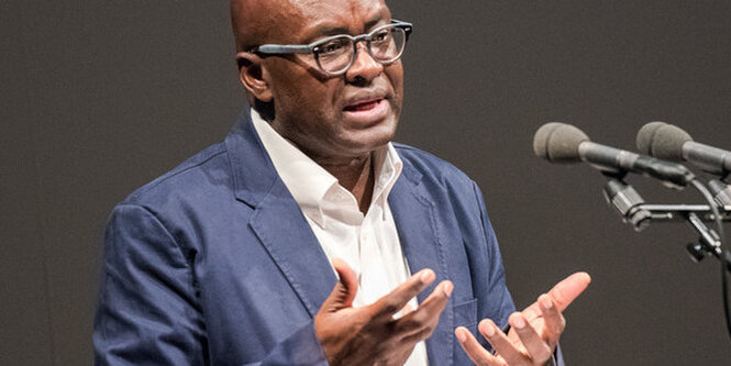 Achille Mbembe steht an zwei Mikrofonen und spricht. Er hebt die Hände zum Gestikulieren. Er trägt eine runde Brille und über seinem weißen Hemd ein dunkelblaues Sakko.