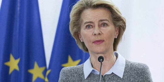Ursula von der Leyen steht an einem Mikrofon. Hinter ihr zwei EU-Flaggen.
