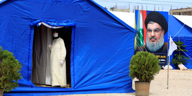 Mitarbeiter in weißer Schutzuniform schaut aus einem blauen Zelt, im Hitnergrund Hizbollah Führer auf einem Plkat