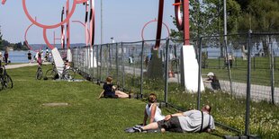 Menschen sitzen auf einer Wiese, die durch einen Zaun getrennt wird und unterhalten sich. Im Hintergrund ein See