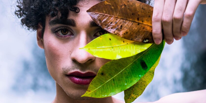 Ein Mann mit dunklen Locken und rotem Lippenstift hält sich kunstvoll Pflanzenblätter vor sein Gesicht