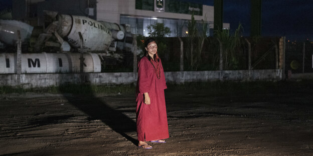 Eine indigene Frau steht nachts vor einem Fabrikgelände.