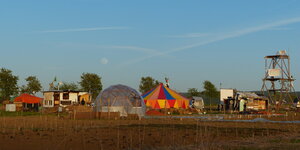 Ein Acker auf dem Hütten, ein rundes Gewächshaus und ein Zirkuszelt stehen