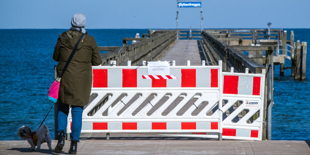 Mit rot-weißer Baustellensicherung abgesperrte Seebrücke, davor eine Frau mit Hund