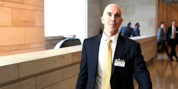 Steve Linick im Anzug und mit gelber Krawatte. Er geht durch ein Foyer, im Hintergrund eine Rolltreppe
