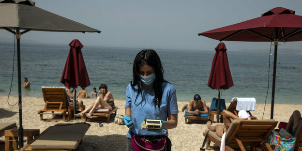 Eine Kellnerin mit Mundschutz am Strand steht zwischen Badeliegen