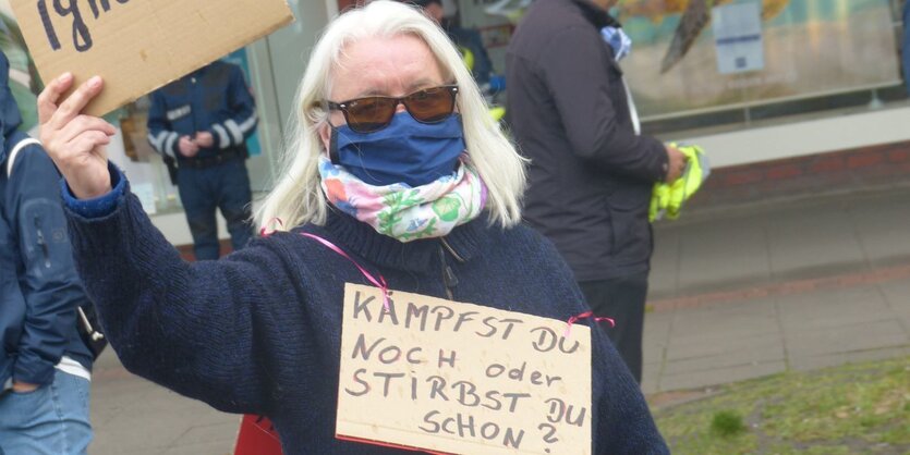 Frau mit Mundschutz und Karton-Plakat um den Hals, Aufschrift: Kämpfst Du noch oder stirbst Du schon?