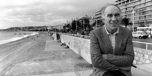 Ein Mann (Michel Piccoli) sitzt im Anzug am Strand und blickt in die Kamera.