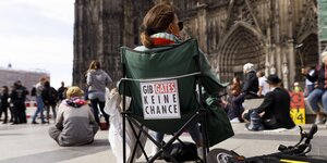 Eine Frau sitzt bei eienr Demo auf einem Campingstuhl, auf dessen Rückseite steht: Gib Gates keine Chance