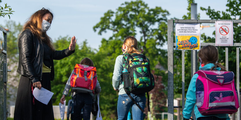 Eine lehrperson steht auf der Linken seite und grüßt die Kinder die durch ein Tor gehen