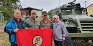 Vier Männer stehen im Freien nebeneinander vor einem Schützenpanzerwagen und halten eine rote Fahne in der Hand, auf der ein Gewehr abgebildet ist