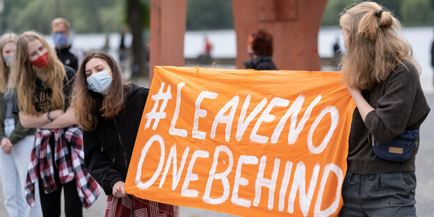 junge Demonstrantinnen mit Mundschutz halten ein Transparent, auf dem steht: "#leavenoonebehind"