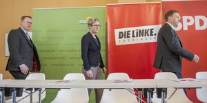 eine Frau und zwei Männer laufen hinter einer Tischreihe entlang, im Hintergrund je ein Plakat der Grünen, der Linken und der SPD